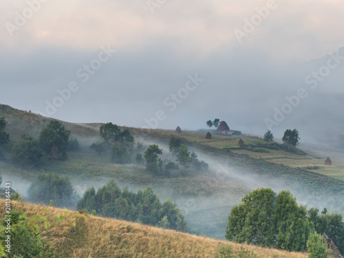 Beautiful mountain landscape with and old house, trees,and haystacks in a foggy morning, Dumesti, Salciua, Apuseni, Romania