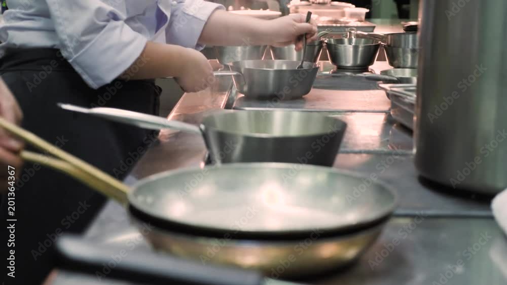 Hands of professional chefs in restaurant or hotel kitchen. Cook during cooking process mixes ingredients of food in frying pan on the stove, use a spoon. A lot of cooking utensils, working in cuisine Hands of professional chefs in restaurant or hotel kitchen. Cook during cooking process mixes ingredients of food in frying pan on the stove, use a spoon. A lot of cooking utensils, working in cuisine