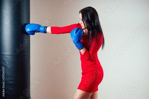 Portrait of a beautiful brunette woman exercising with boxing gloves on her hands in the gym. athlete with make-up red lipstick and dress boxing in the gym. the concept of women's power and feminism © yurakrasil