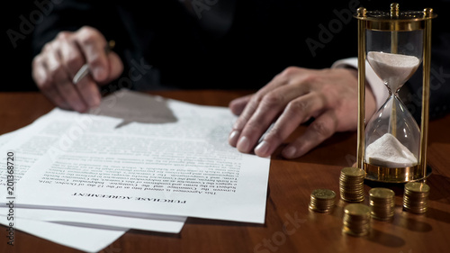 Businessman signing purchase agreement as time flows with sand in clock, closeup