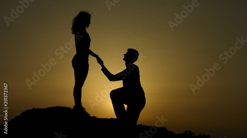 Male silhouette making proposal to woman on top of mountain at sunset time