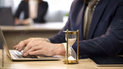 Fotografia Businessman typing on laptop at desk, hourglass trickling, deadline approaching