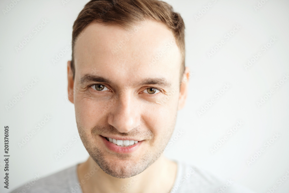 Naklejka premium Handsome young man with a bristle smiling teeth. Portrait of a guy in a gray shirt on a light background.