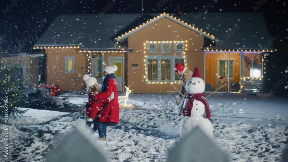 On the Happy Winter Evening Young Father, Mother and Cute Little Daughter Play in Snowballs, Running around the Snowman They've Built in the Backyard of Their Idyllic House Decorated with Garlands. 