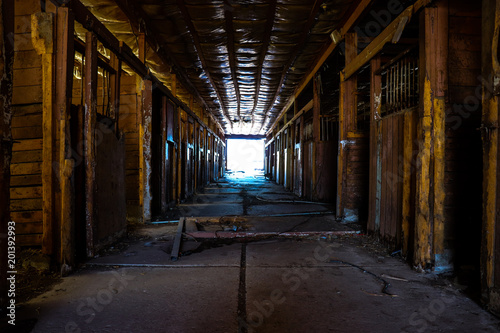 Empty horse stalls within an abandoned dilapidated barn with clean line shining through the open barn doors at the end of the room