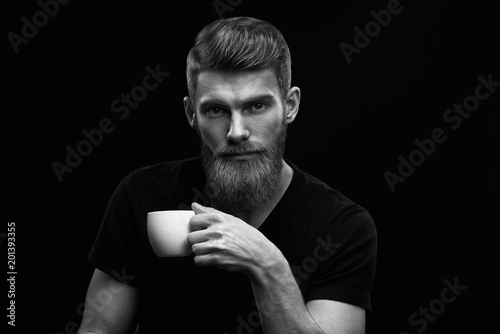 Black and white dramatic light studio shot of bearded stylish young man with cup of coffee in hand Handsome man having coffee look at camera Male taking rest and drinking fresh morning espresso .