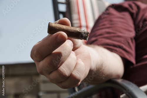 Dirty hand holding a burning cigar, with a blurred man in a red shirt sitting on a chair in the background