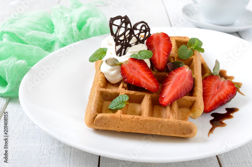 Whole wheat Belgian waffles with whipped cream, freshly chopped strawberries, mint leaves and chocolate on white wooden background. Homemade healthy breakfast.