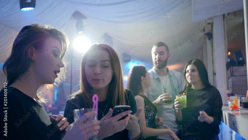 girlfriends gossip at the bar girl with an alcoholic beverage looks at the screen of mobile phone behind the bar counter on background of lumiere