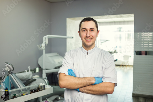 Male dentist standing with his hands crossed over medical office background. Healthcare, profession, stomatology and medicine concept