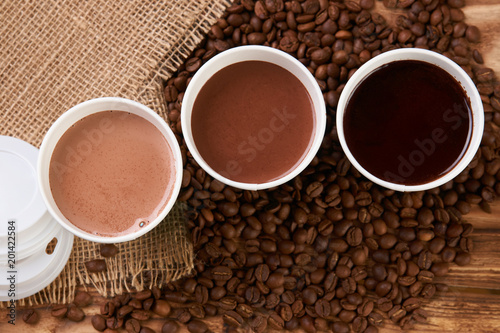 Three take away green paper cups with coffee beans and hot chocolate drink on wooden background, top view, close-up