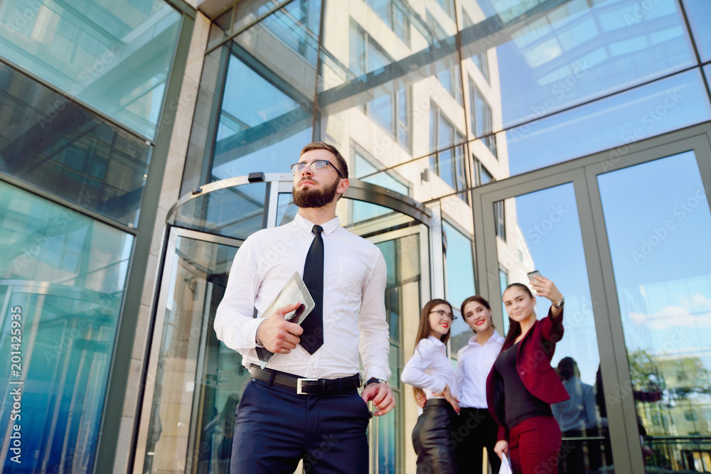 A young male manager with a tablet in his hands against the background of the girls. Office staff in the background of a multi-storey glass building. Lifestyle. Businessman A young male manager with a tablet in his hands against the background of the girls. Office staff in the background of a multi-storey glass building. Lifestyle. Businessman