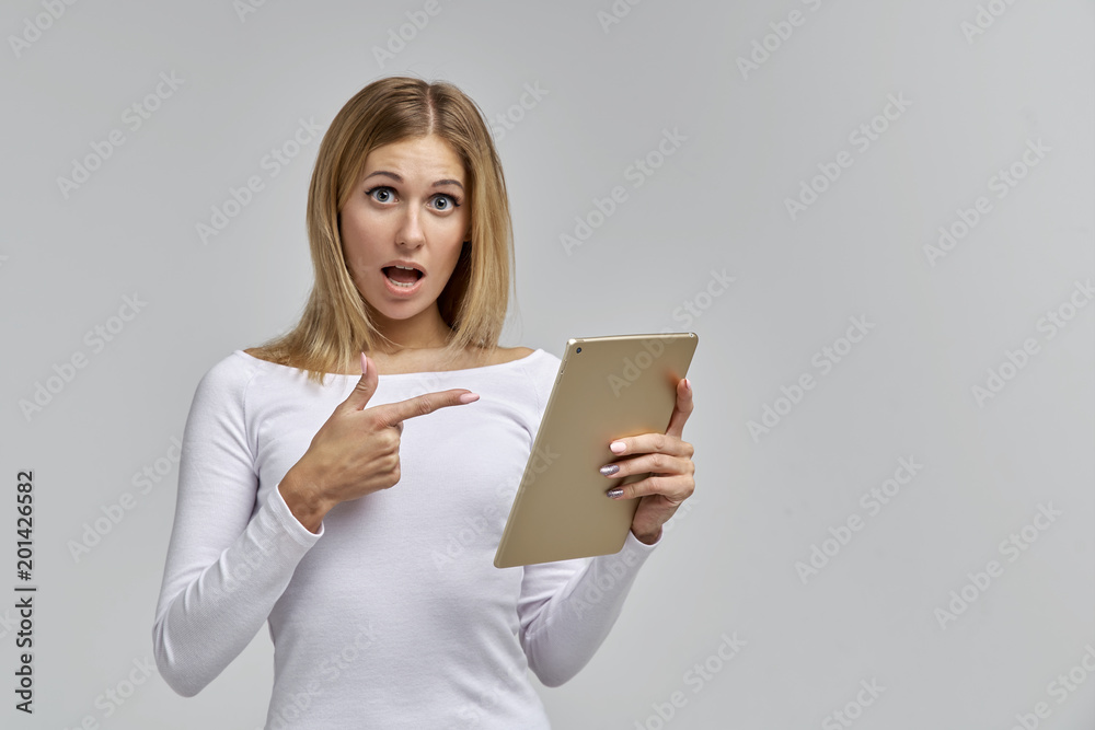 beautiful slender young blonde woman shows her finger on a tablet in her hand and opens her eyes and mouth wide beautiful slender young blonde woman shows her finger on a tablet in her hand and opens her eyes and mouth wide