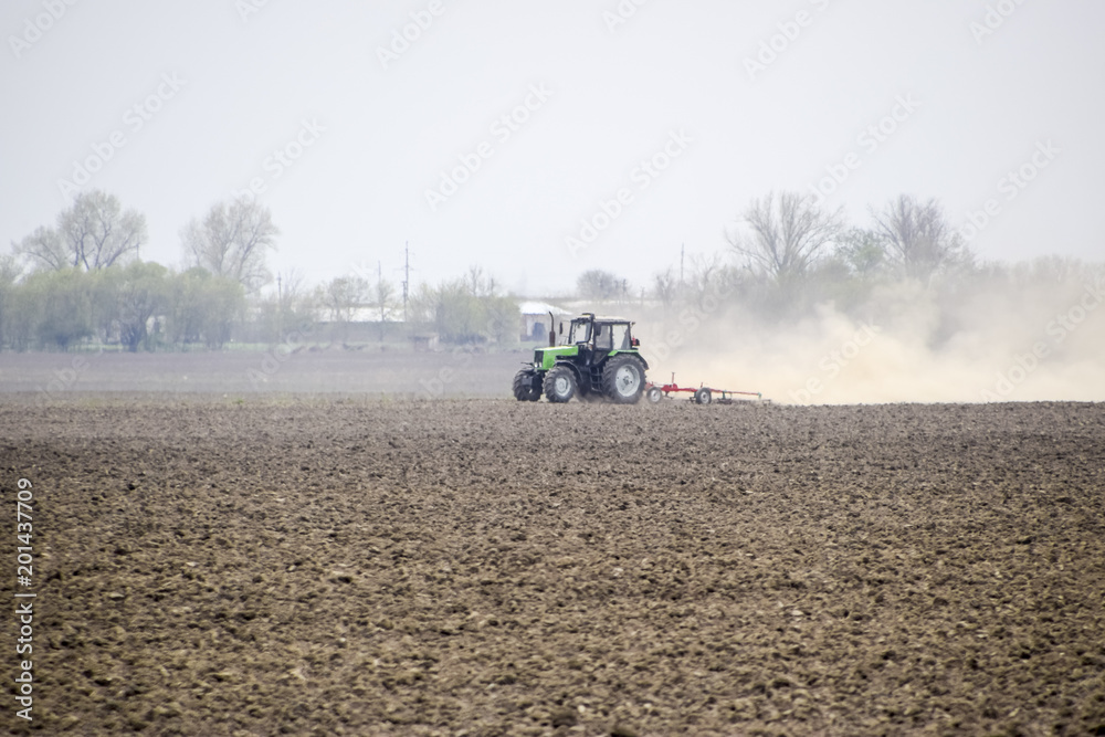Naklejka premium The tractor harrows the soil on the field and creates a cloud of dust behind it
