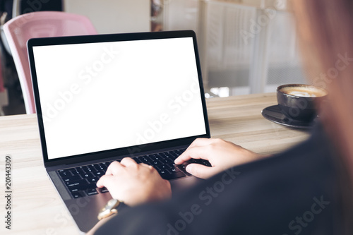 Mockup image of a businesswoman using and typing on laptop with blank white desktop screen with coffee cup on wooden table in office