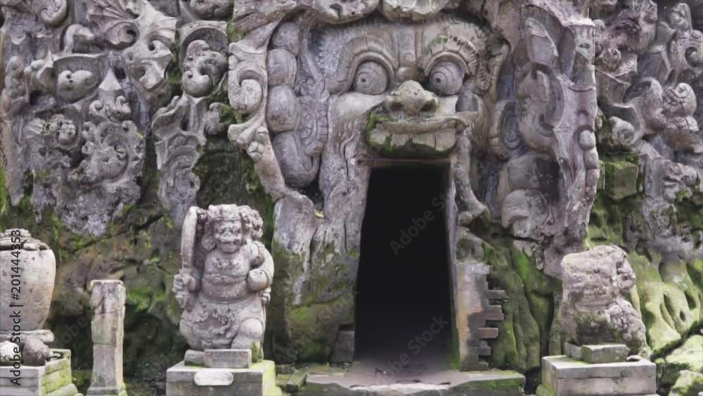 Enterance to the elephant cave the in the wild tropical jungle, detailing the weathered surface with white fungus and moss. Lush vegetation and Balinese temple on the background. Slow tracking shot Enterance to the elephant cave the in the wild tropical jungle, detailing the weathered surface with white fungus and moss. Lush vegetation and Balinese temple on the background. Slow tracking shot