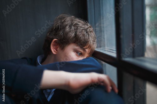 Pensive sad boy teenager with blue eyes in a blue shirt and jeans sitting at the window and closes his face with his hands. photo