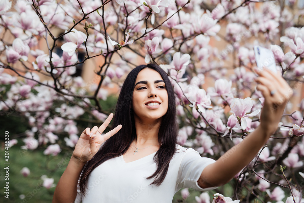 Beautiful stylish caucasian woman making selfie in blossom magnolia garden.