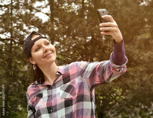 Beautiful woman wearing checked shirt and baseball cap taking selfie in the city park. Selective focus. © zdravinjo