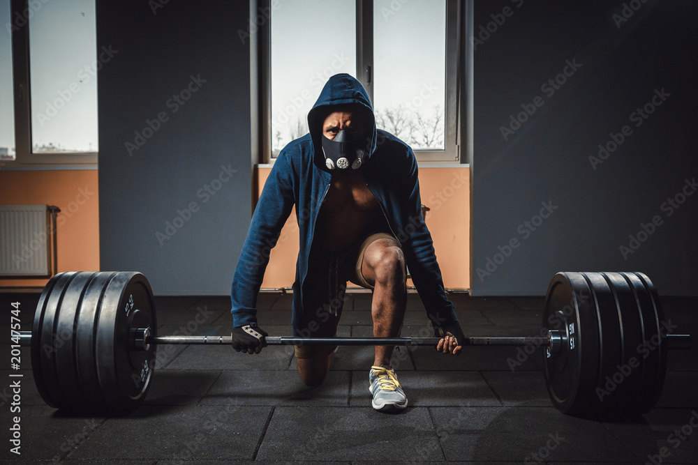 athletic man in jacket with a hood waiting and preparing before lifting heavy barbell looking at camera. fitness sport training gym concept. deadlift with barbell in mask. bent over the barbell Stock