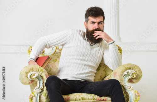 Connoisseur of literature concept. Macho spends leisure with book. Connoisseur, on thoughtful face enjoy literature. Man with beard and mustache sits on armchair and reading, white wall background.