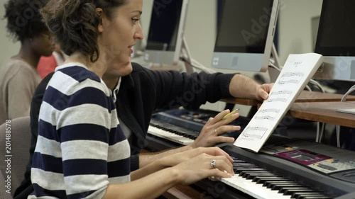 Woman learning to play electric keyboard in college music lesson with tutor helping.