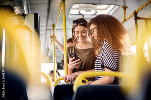 Two cheerful pretty young women are standing in a bus and looking at the phone and smiling while waiting for a bus to take them to their destination. photo