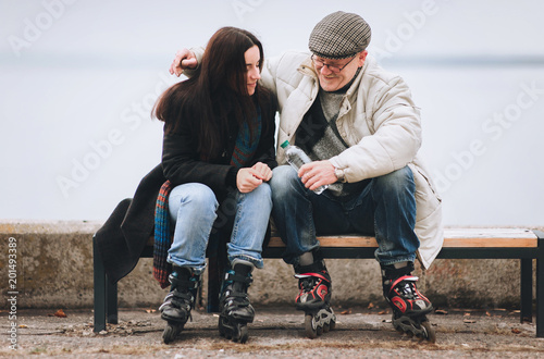 Father and daughter on roller skates sit on a bench. They smile cheerfully. The man in his hands has a bottle of water. Active old people.