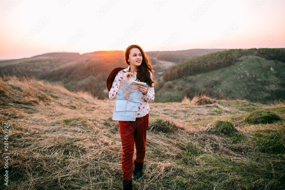 Stylish traveler woman with hat holding map on top of mountains, emotional happy moment, travel concept, space for text. Girl  walking along the grass at the top of the mountain in sunset