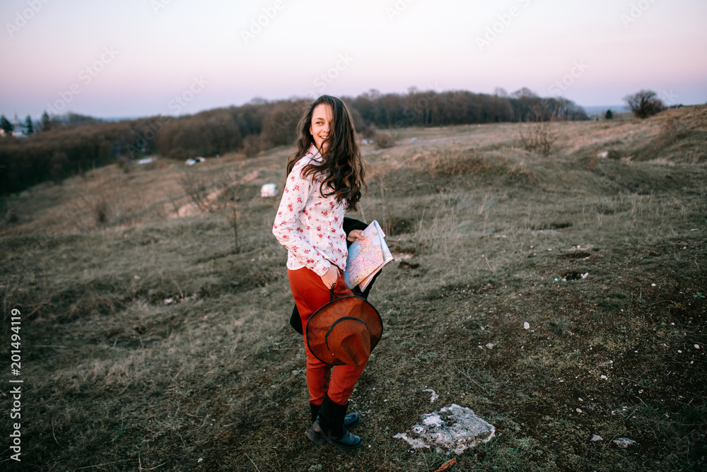 Stylish traveler woman with hat holding map on top of mountains, emotional happy moment, travel concept, space for text. Girl  walking along the grass at the top of the mountain in sunset