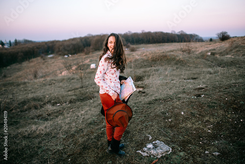 Stylish traveler woman with hat holding map on top of mountains, emotional happy moment, travel concept, space for text. Girl  walking along the grass at the top of the mountain in sunset