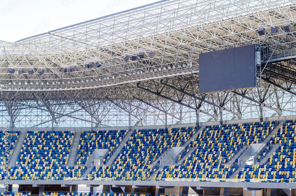 Fototapeta premium Siedzący kibice kolorowe plastikowe krzesła na tle stadionu piłkarskiego. Pusty stadion boisko do piłki nożnej zielona trawa na arenie lekkoatletycznej piłki nożnej.