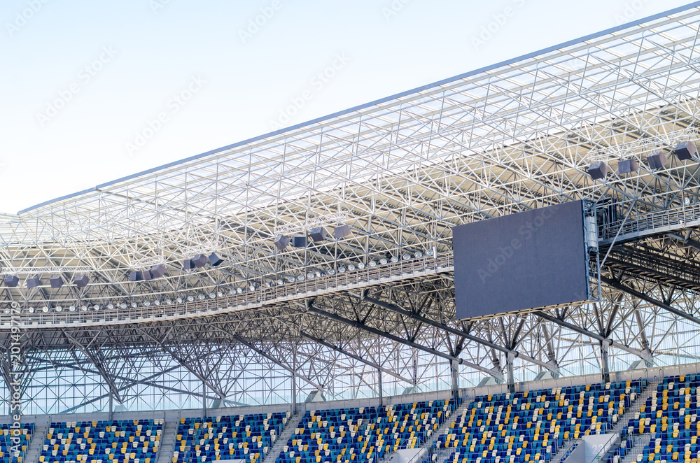 Fototapeta premium Siedzący kibice kolorowe plastikowe krzesła na tle stadionu piłkarskiego. Pusty stadion boisko do piłki nożnej zielona trawa na arenie lekkoatletycznej piłki nożnej.