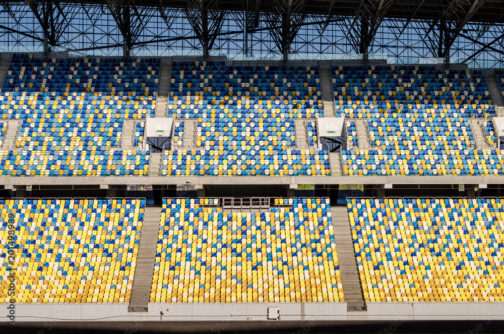 Obraz premium Niewyraźne siedzi fanów kolorowe plastikowe krzesła na tle stadionu piłkarskiego. Puste boisko piłkarskie stadionu zielona trawa na arenie lekkoatletycznej piłki nożnej.
