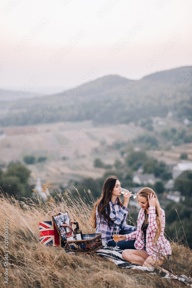 Two beautiful girls in colored shirts sitting on the grass on the hillside. Watch the sunset, drinking wine, hug, happy. picnic