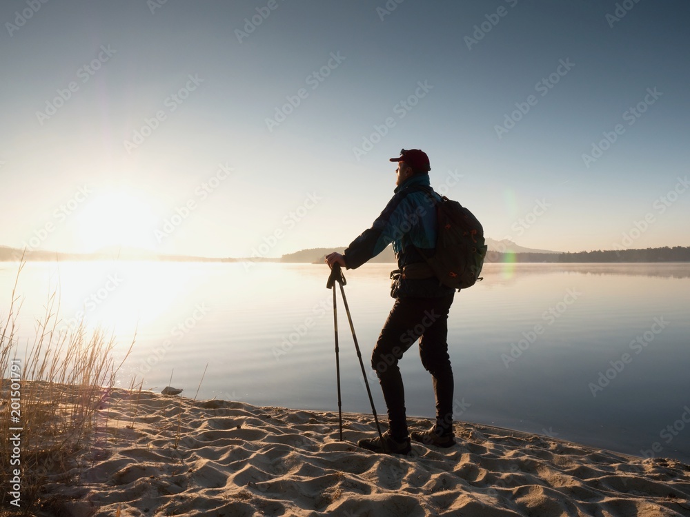 Man tourist with a backpack and trekking poles standing on sandy beach. Hiker enjoy