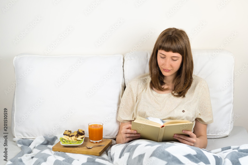 Young woman smiles and reads book in bed and has breakfast. Female person lying in clean white minimalistic bed at home or hospital with healthy vegan diet breakfast and juice glass
