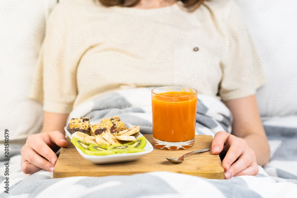 Fresh raw vegetarian breakfast in bed. Morning vegan meals of cereal candy, banana, kiwi and fresh vegetable or fruit juice in female hands in bedroom