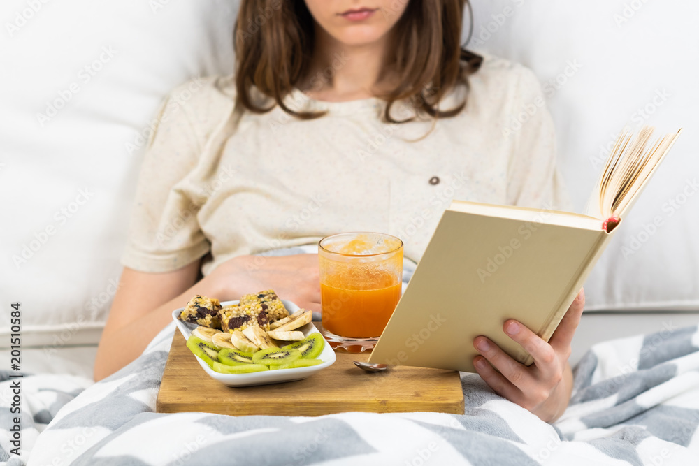 Reading book in bed and having breakfast. Female person lying in clean white minimalistic bed at home or hospital with healthy vegan diet breakfast and juice glass Reading book in bed and having breakfast. Female person lying in clean white minimalistic bed at home or hospital with healthy vegan diet breakfast and juice glass