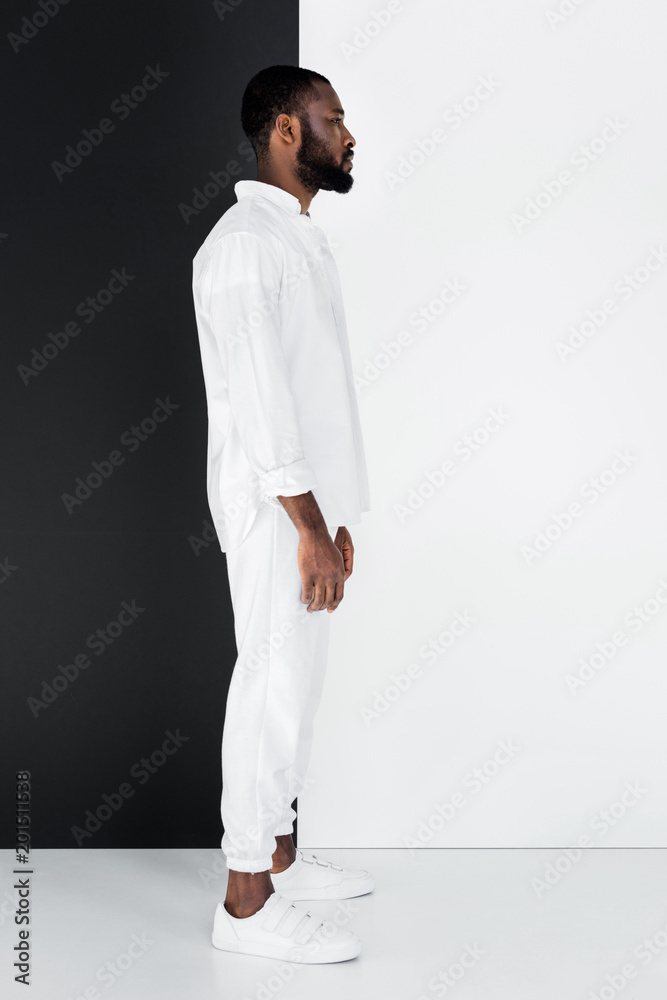 side view of stylish african american man standing in white clothes near black and white wall side view of stylish african american man standing in white clothes near black and white wall