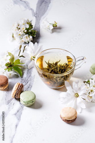 Glass cup of hot green tea with french dessert macaroons, spring flowers white magnolia and cherry blooming branches over white marble texture background.