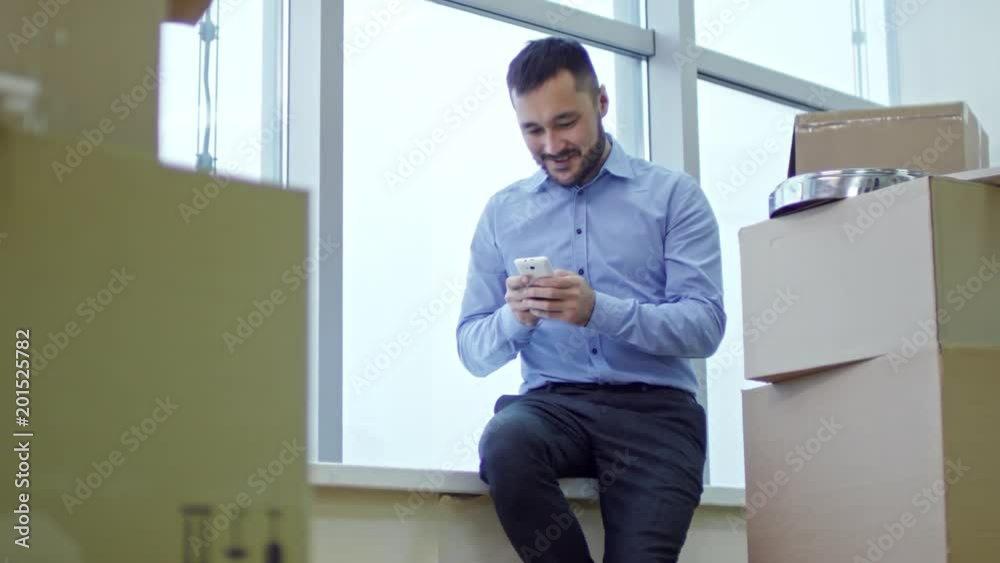 Tracking shot of asian businessman sitting on window sill in new office with stacks of cardboard boxes, smiling and messaging on smartphone Tracking shot of asian businessman sitting on window sill in new office with stacks of cardboard boxes, smiling and messaging on smartphone