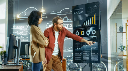 Female Developer and Male Statistician Use Interactive Whiteboard Presentation Screen to Look at Charts, Graphs and Growth Statistics. They Work in the Stylish Creative Office.