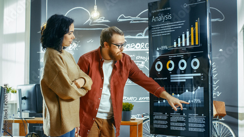 Female Developer and Male Statistician Use Interactive Whiteboard Presentation Touchscreen to Look at Charts, Graphs and Growth Statistics. They Work in the Stylish Creative Office. photo