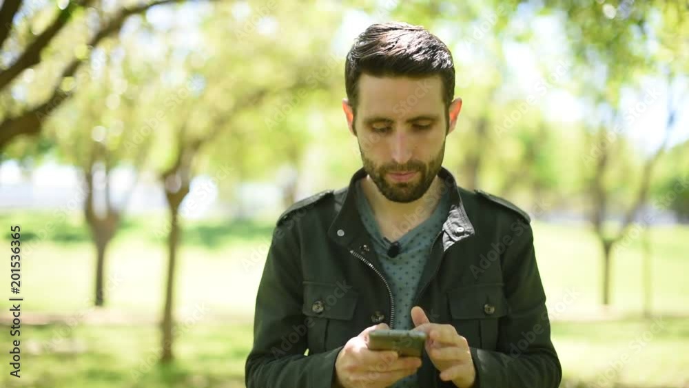 Hombre joven moreno con chaqueta verde mirando el móvil en un parque/bosque