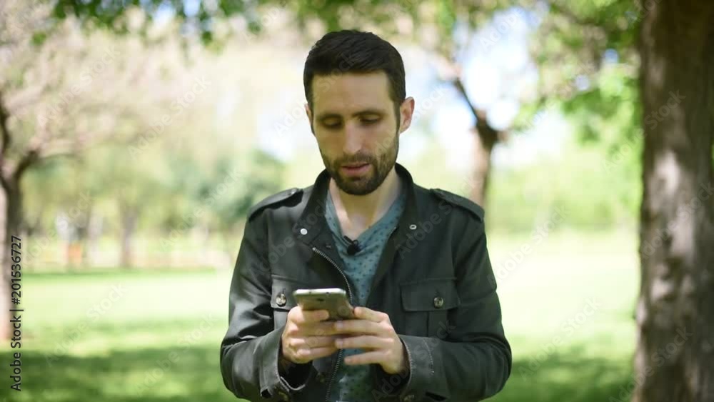 Hombre joven moreno con chaqueta verde mirando el móvil en un parque/bosque
