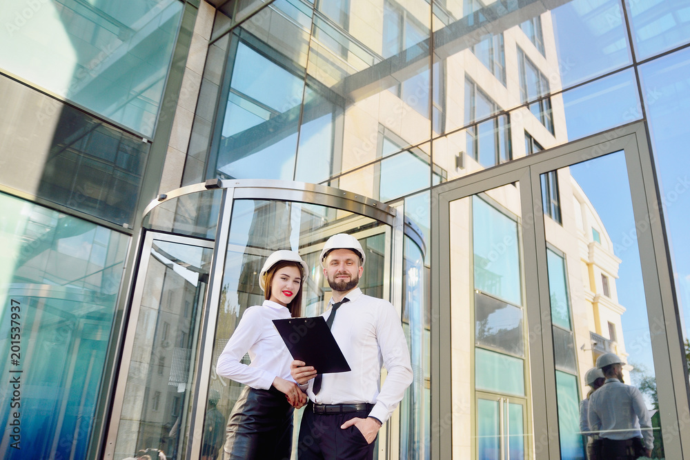 A girl with a man in construction white helmets and white shirts with drawings in hands against a glass office building. Engineers. Planning of construction A girl with a man in construction white helmets and white shirts with drawings in hands against a glass office building. Engineers. Planning of construction