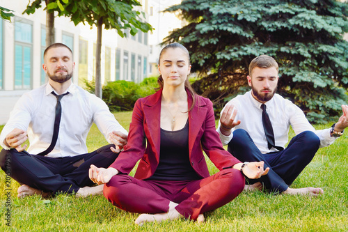 Office yoga. Three young employees in a lotus pose are sitting on green grass and meditate. Recreation of office staff. Healthy lifestyle. Guys and the girl
