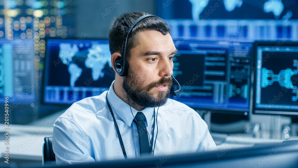 System Control Room Dispatcher Talks into Headset. He Controls Correct Work of the Facility. In the Background Multiple Monitors Show Technical Data. System Control Room Dispatcher Talks into Headset. He Controls Correct Work of the Facility. In the Background Multiple Monitors Show Technical Data.