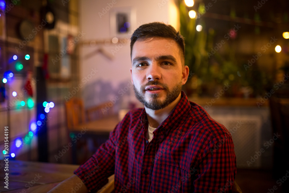 Portrait of a young handsome Caucasian man with a beard and toothy smile in a red checkered shirt sitting beside a laptop at a wooden table. In the evening at the coffee shop Portrait of a young handsome Caucasian man with a beard and toothy smile in a red checkered shirt sitting beside a laptop at a wooden table. In the evening at the coffee shop